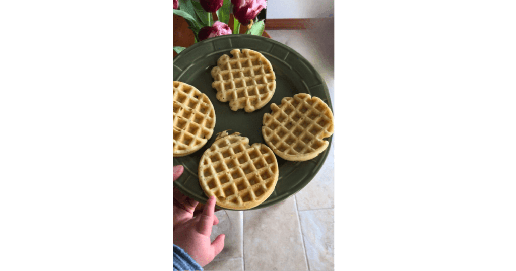 Four waffles on a green plate with a baby hand and tulips in the background