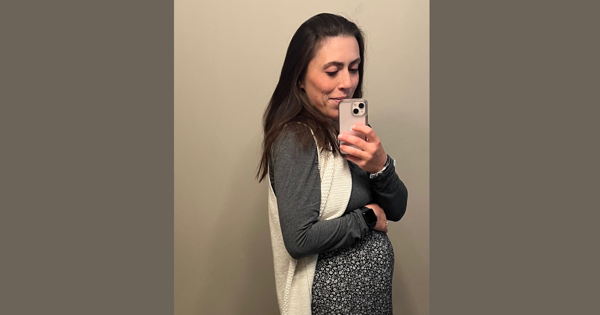 Girl with brown hair and pink iPhone taking a picture with a white shawl, grey shirt and black/white floral skirt