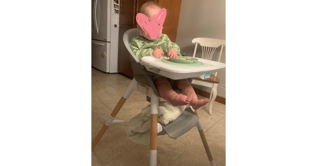 Baby in white and grey high chair wearing green bib.