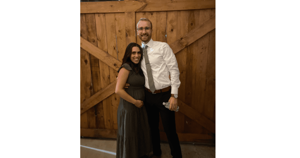 Husband in dress pants and white shirt with green tie. Wife in green dress holding stomach. Barn doors with criss cross in background.