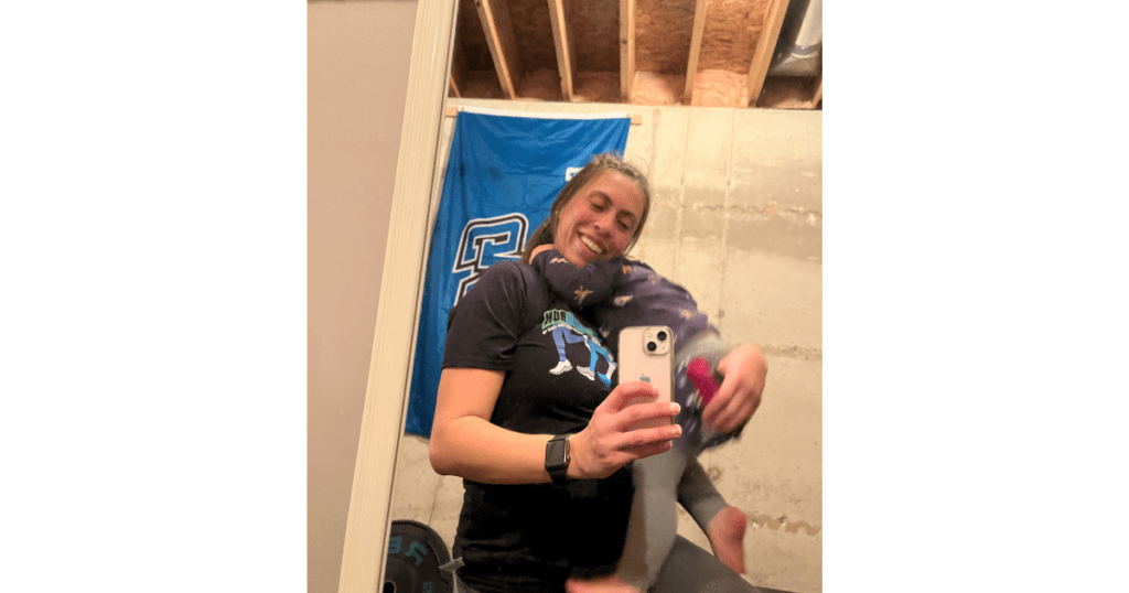 Mom kneeling while holding daughter. Snapping a picture in front of a blue flag in basement with exposed ceilings.