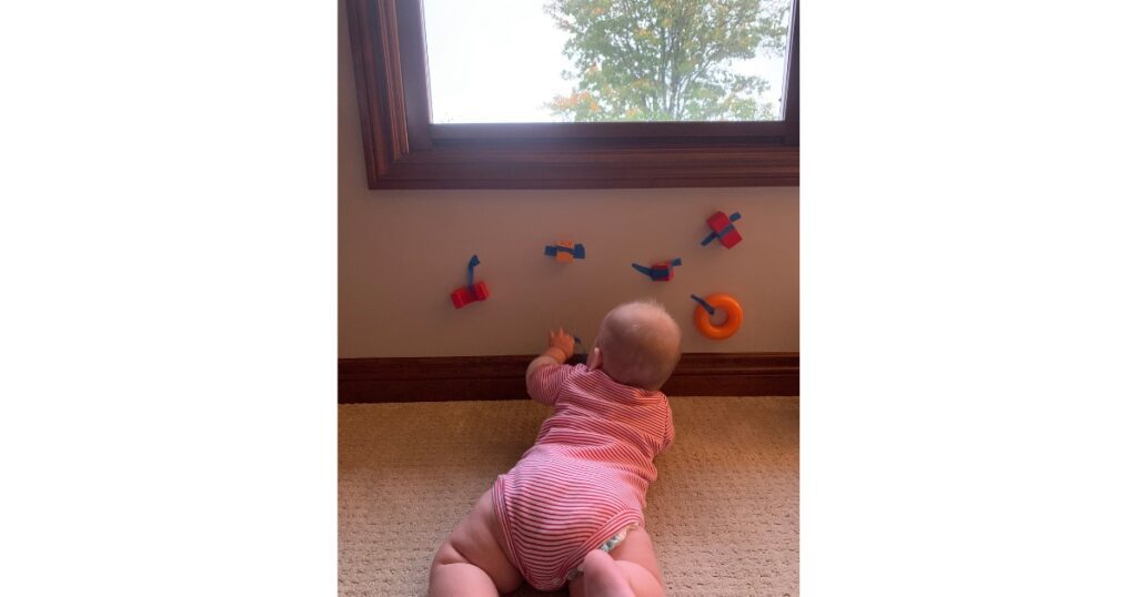Baby in a pink onesie laying on their stomach and pulling toys taped to the wall.