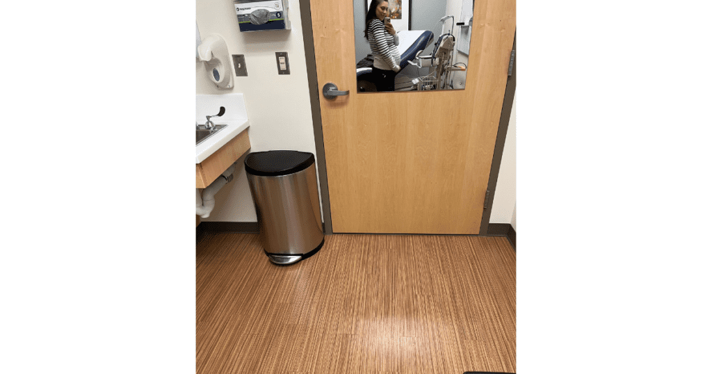 Girl in doctors office taking a picture in a mirror. Gray trash can in corner. Medical equipment next to bed.