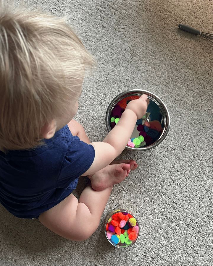 Child with blonde hair playing with pom poms in a bowl.