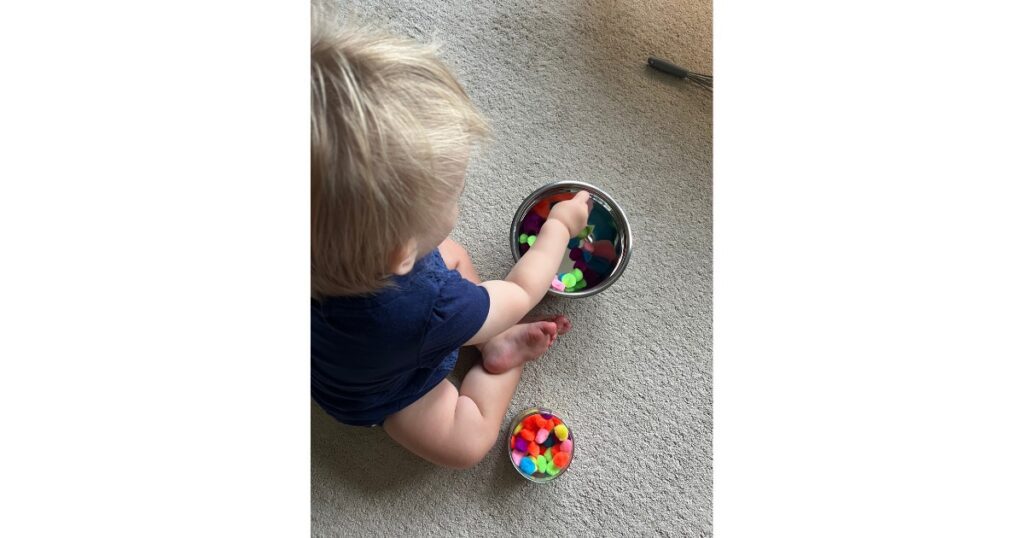 Child with blonde hair playing with a bowl of pom poms.