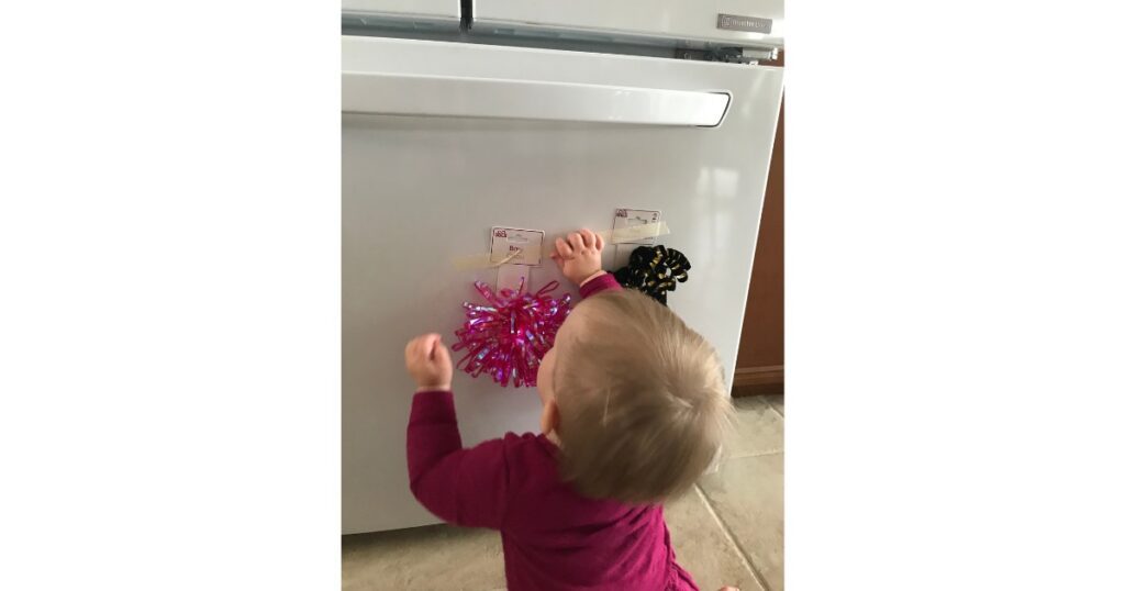 Toddler with blonde hair and pink shirt playing with ribbons on refrigerator