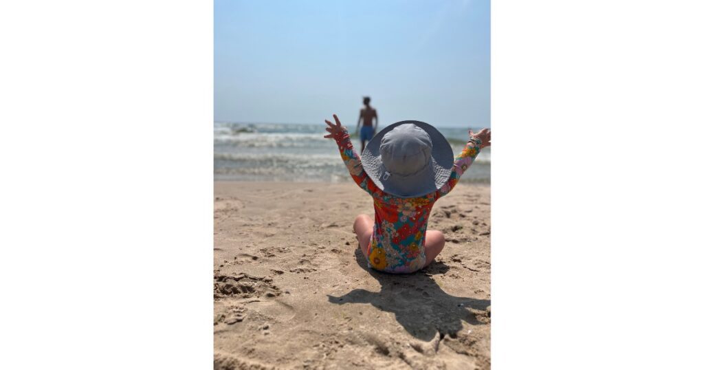 Child at beach in flower one piece bathing suit. Hands up in air. No clouds in sky.