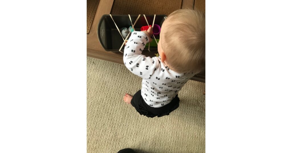Toddler standing up next to a table with blonde hair and bow onesie. Pulling toys out of a bread pan with rubber bands.