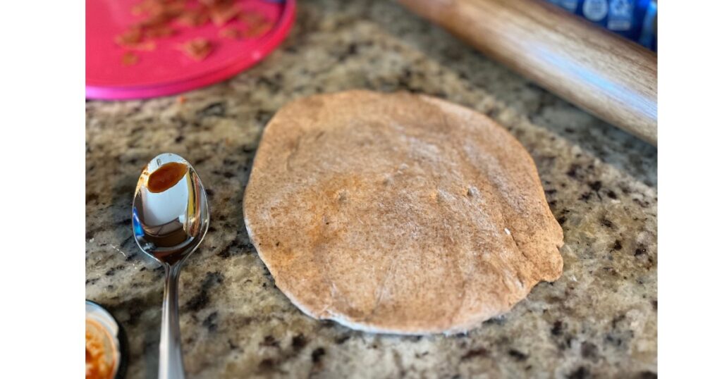 whole wheat pizza crust next to spoon on counter. rolling pin in background