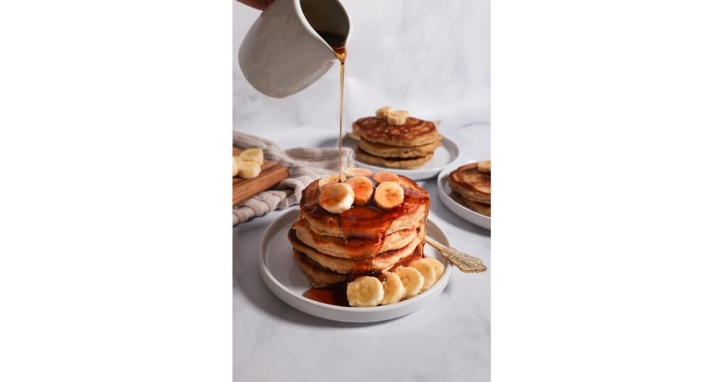 maple syrup coming out of a container and being put on pancake stack with sliced bananas