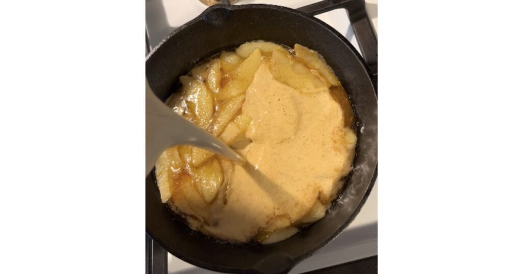 pouring dutch baby batter into a cast iron skillet over apples